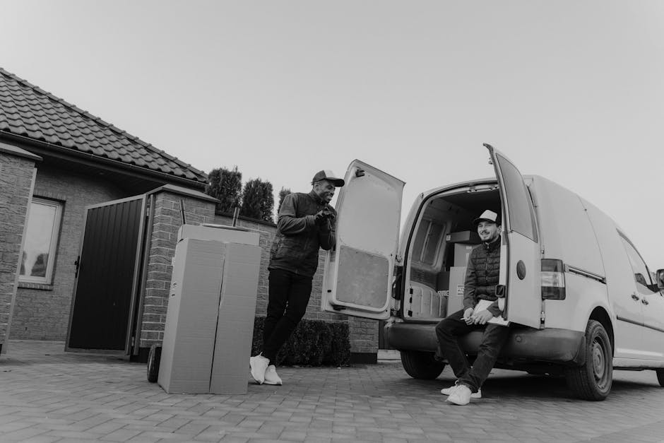 Two men are engaged in a home relocation task, situated outdoors on a driveway next to a brick house with a tiled roof. One man, standing beside the open rear door of a white delivery van, is using a mobile phone while wearing casual clothing, including a hat and a jacket. The other man, seated on the edge of the van's open rear platform, is dressed in a hoodie, jacket, trousers, and white sneakers, smiling at the camera. Several cardboard boxes, some covered with plastic wrap, are placed on the driveway, with additional packing materials visible nearby. The van's interior contains more boxes and packing related to furniture transport and moving logistics. The setting suggests a loading and unloading process involved in a house removal, with the van positioned on a paved surface close to the home. Man and Van Dulwich offers professional removals and relocation services, supporting efficient packing and furniture transport.