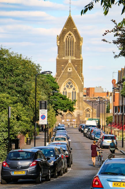 A residential street scene featuring a row of parked cars along the pavement, with a woman wearing a red coat and a person riding a bicycle nearby. The street is lined with trees and traditional brick buildings, leading towards a prominent, tall stone church with gothic-style windows and a pointed spire in the background. Street lamps and signage are visible, indicating parking restrictions and zones. The overall environment appears calm, with soft daylight illuminating the scene. This setting exemplifies a typical urban area where a house removal or furniture transport process could take place, possibly involving the loading or unloading of moving boxes and furniture near the curb as part of home relocation services provided by companies like Man and Van Dulwich.