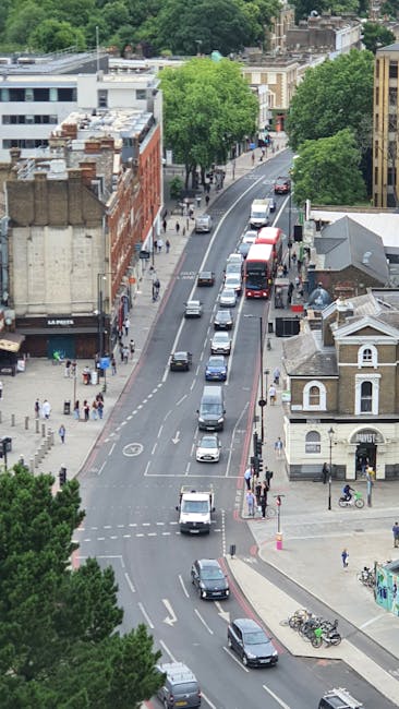 An aerial view of a busy street in Dulwich showing multiple vehicles including cars and a double-decker bus moving along the road, with pedestrians walking on the sidewalks, some waiting to cross at designated crossings, and a variety of buildings lining the street such as shops, residential houses, and trees providing greenery. The scene captures the typical urban environment involved in household relocations and furniture transport, with parked bicycles near a corner, and a moving van or truck not visible but implied as part of a relocation process associated with Dulwich removals. The lighting suggests daytime with natural daylight illuminating the scene, highlighting the organized flow of traffic and pedestrian activity typical during home relocation and moving logistics handled professionally by companies like Man and Van Dulwich.