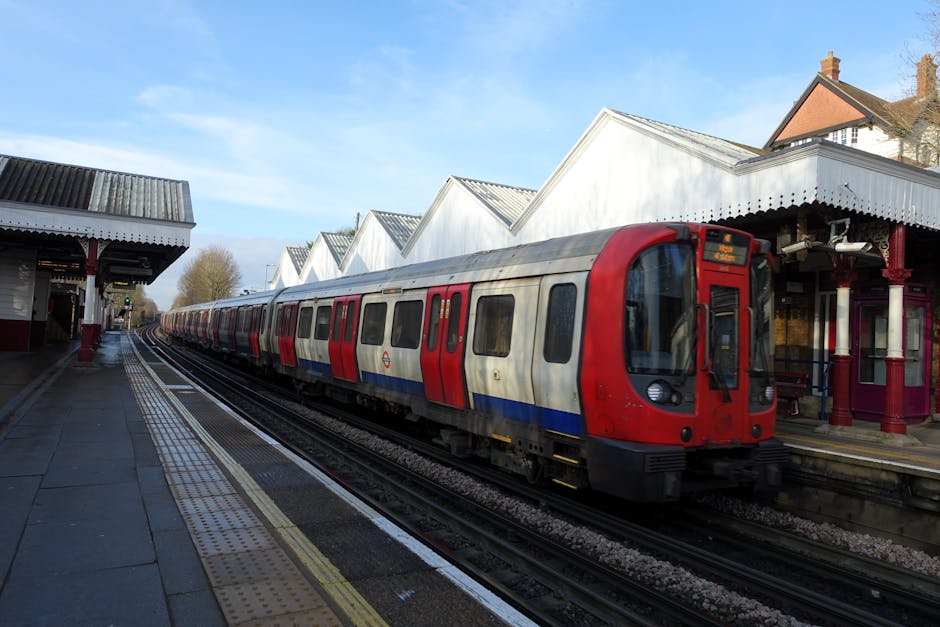 A modern London Underground train with a red front and white, blue, and red livery is stationary at North Dulwich station platform on a clear day. The platform features tactile paving strips along the