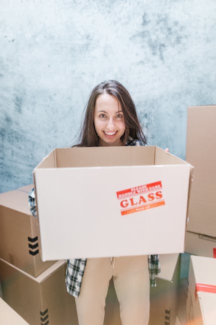 A woman with long brown hair, smiling and wearing a checkered shirt, stands indoors in front of a plain light blue wall, holding an empty cardboard box labeled 'GLASS' in red text. Around her are seve